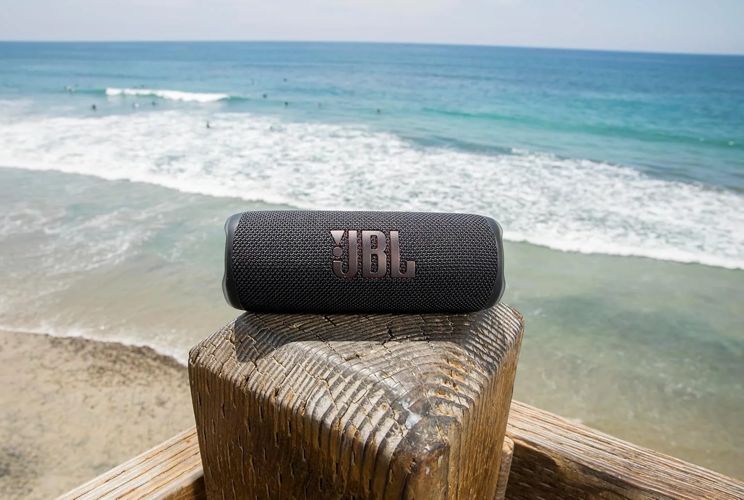 Black JBL Flip 6 speaker on a railing above the beach